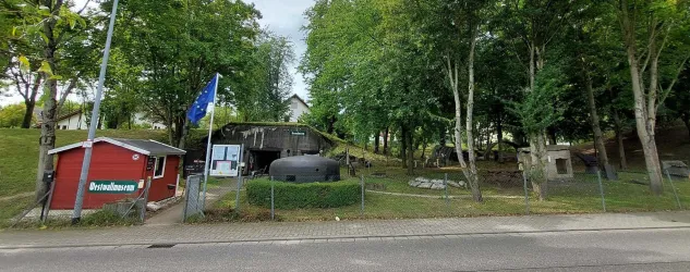 Eine kleine Anlage mit mehreren Gebäuden und einem großen Wasserbehälter in einer bewaldeten Umgebung. Im Vordergrund steht ein rotes Häuschen mit einer Flagge.