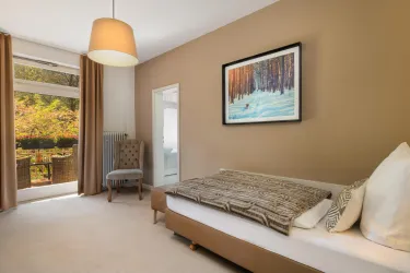 Ein helles Schlafzimmer mit einem Einzelbett und einem gemütlichen Sessel. Große Fenster bieten einen Blick auf den Garten.