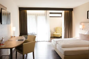 Ein modernes Hotelzimmer mit einem Doppelbett und einem Schreibtisch. Große Fenster lassen viel Tageslicht herein und bieten einen gemütlichen Sitzbereich.