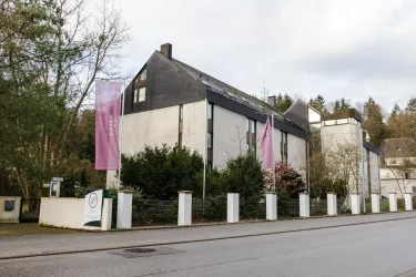 Ein modernes Gebäude mit einer markanten Architektur und großen Fenstern. Blühende Pflanzen und Straßenbanner verleihen der Umgebung einen einladenden Eindruck.