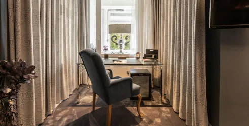 Ein modernes Arbeitszimmer mit einem Schreibtisch und einem bequemen Stuhl. Große Vorhänge und ein Fenster sorgen für viel Tageslicht.