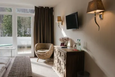 Ein gemütliches Zimmer mit einem bequemen Stuhl und einem Fernseher an der Wand. Das Fenster bietet einen Blick auf einen Außenbereich mit Pflanzen.