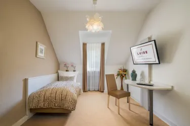 Ein gemütliches Zimmer mit einem Einzelbett, einem Schreibtisch und einem Stuhl. Die Wände sind in einem warmen Braunton gestrichen und das Zimmer hat große Fenster mit Vorhängen.