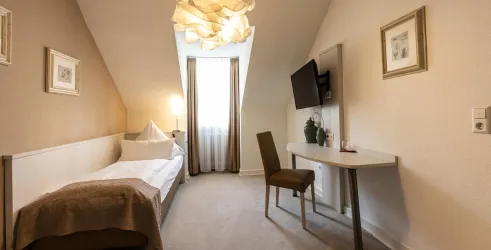 Ein gemütliches Zimmer mit einem Einzelbett, einem Schreibtisch und einem Fernseher. Das Licht von der Lampe und das Fenster sorgen für eine helle und freundliche Atmosphäre.