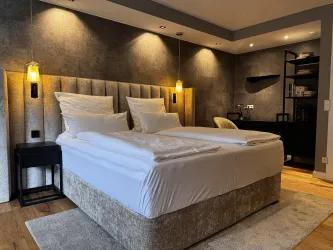 Ein modernes Hotelzimmer mit einem großen, bequemen Bett und eleganter Beleuchtung. Die Wände sind in einem dunklen Ton gehalten und es gibt stilvolle Möbel.