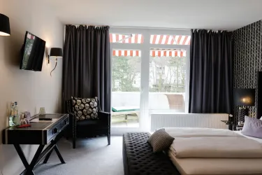Ein modernes Hotelzimmer mit einem großen Fenster und einer Terrassentür. Es gibt ein Bett, einen Sessel und einen Schreibtisch, alles in stilvollen Farben.