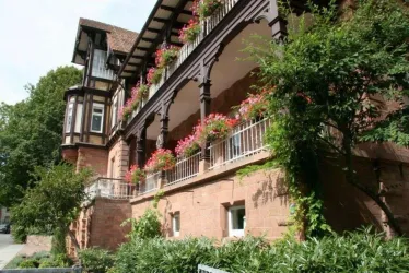 Ein historisches Gebäude mit einem schönen Balkon, der mit blühenden Pflanzen geschmückt ist. Die Fassade ist aus rotem Stein und von Bäumen umgeben.