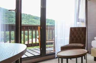 Ein gemütlicher Innenraum mit einem bequemen Sessel und einem runden Tisch. Im Hintergrund ist ein Balkon mit Blick auf grüne Hügel zu sehen.