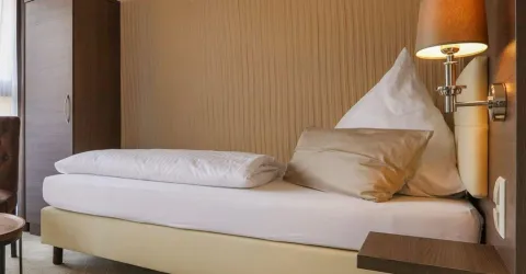 Ein einfaches Einzelzimmer mit einem Bett, einem Kissen und einer Lampe. Die Wände sind beige und der Raum wirkt gemütlich.