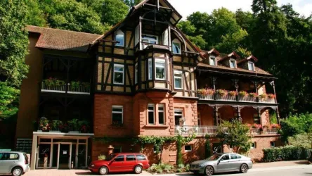 Ein charmantes Gebäude im traditionellen Stil, umgeben von Bäumen. Auf den Balkonen blühen Pflanzen, und vor dem Haus stehen einige Autos.