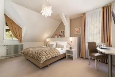 Ein elegantes Zimmer mit einem Doppelbett und weichem Teppichboden. Es gibt einen kleinen Tisch mit Stühlen und viel Tageslicht durch die Fenster.