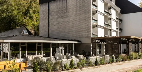 Ein modernes Gebäude mit mehreren Etagen und einem angrenzenden Terrassenbereich. Die Umgebung ist grün und einladend gestaltet.