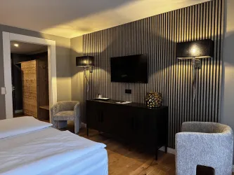 Ein modernes Hotelzimmer mit einem Doppelbett und minimalistischer Einrichtung. Die Wände sind dezent gestreift und es gibt einen Fernseher sowie bequeme Sessel.