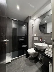 Ein modernes Badezimmer mit einer Dusche und einem runden Spiegel. Die Wände sind in Grautönen gehalten, und es gibt frische Handtücher auf einem Regal.