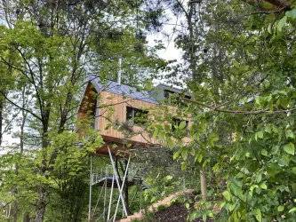 Ein modernes Baumhaus umgeben von grünen Bäumen. Das Haus hat eine Holzfassade und steht auf Stelzen.