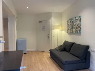 Ein einfaches Wohnzimmer mit einem grauen Sofa und einem kleinen Tisch. An der Wand hängt ein buntes Kunstwerk, und es gibt einen Kühlschrank in der Ecke.