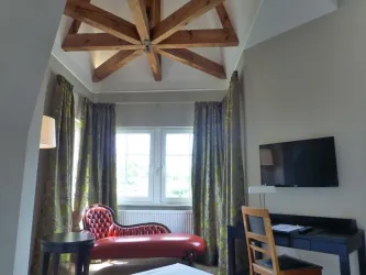 Ein gemütlicher Wohnraum mit einem roten Sofa und großen Fenstern. An der Wand ist ein Fernseher angebracht, und die Decke hat sichtbare Holzbalken.