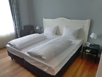 Ein gemütliches Schlafzimmer mit einem Doppelbett und zwei großen Kissen. Die Wände sind in einem hellen Grau gestrichen, und es gibt einen Sessel sowie Tischlampen.