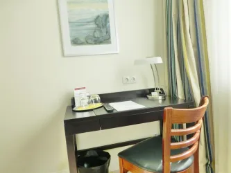 Ein einfacher Schreibtisch mit einem Stuhl in einem hellen Raum. Auf dem Tisch befinden sich Wasserflaschen, Notizen und eine Lampe.