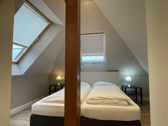 Ein gemütliches Schlafzimmer mit zwei Einzelbetten und schrägen Decken. Es gibt kleine Nachttische und Fenster, die Tageslicht hereinlassen.