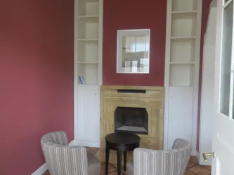 Ein gemütlicher Raum mit einer rosa Wand, zwei bequemen Stühlen und einem kleinen Tisch. Der Kamin in der Mitte verleiht dem Raum eine einladende Atmosphäre.