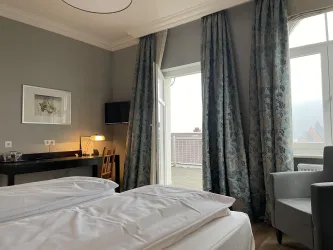 Ein modernes Hotelzimmer mit zwei Betten und stilvollen Vorhängen. Es gibt einen Schreibtisch und einen gemütlichen Sessel neben einem Fenster, das auf einen Balkon führt.