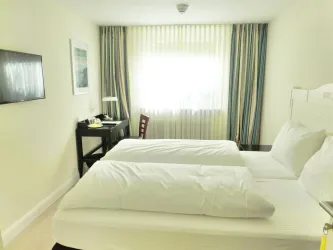 Ein helles Hotelzimmer mit zwei Einzelbetten und einem Schreibtisch. Große Fenster sorgen für viel Tageslicht.