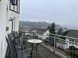 Ein Balkon mit mehreren Tischen und Stühlen. Im Hintergrund sind Hügel und Häuser in einer nebeligen Atmosphäre zu sehen.