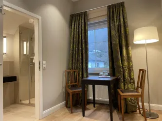 Ein gemütliches Zimmer mit zwei Stühlen und einem Tisch. Der Raum hat große Fenster mit Vorhängen und eine Dusche im Hintergrund.