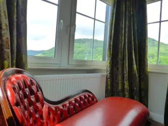 Ein rotes, gepolstertes Sofa steht vor einem großen Fenster mit Blick auf grüne Hügel. Die Vorhänge sind dekorativ und lassen viel Licht herein.