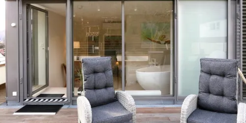 Eine moderne Terrasse mit zwei bequemen Stühlen und einer großen Glastür, die zum Innenraum führt. Im Hintergrund ist ein Badezimmer mit einer eleganten Badewanne zu sehen.