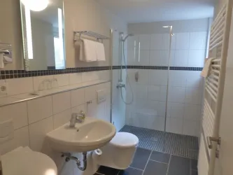 Ein modernes Badezimmer mit einer Glasdusche und hellen Fliesen. Es gibt ein Waschbecken, eine Toilette und Handtücher an der Wand.