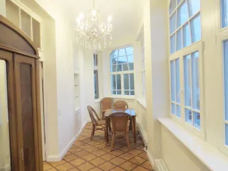 Ein heller Flur mit großen Fenstern und einem eleganten Kronleuchter. In der Mitte steht ein Tisch mit Stühlen aus Rattan.