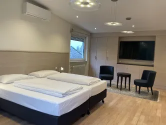 Ein modernes Schlafzimmer mit einem Doppelbett und zwei Stühlen. Große Fenster sorgen für viel Tageslicht.