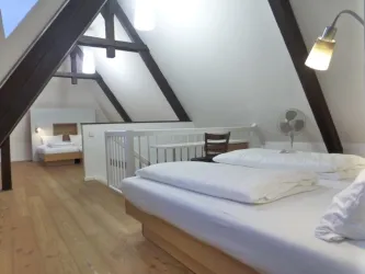 Ein gemütliches Dachzimmer mit Holzfußboden, zwei Betten und einem kleinen Tisch. Große Fenster sorgen für viel Tageslicht.