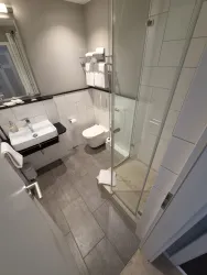 Ein modernes Badezimmer mit einer Dusche und einem Waschbecken. Die Wände sind hellgrau und es gibt weiße Handtücher.