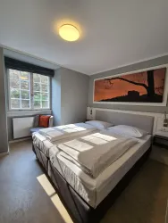 Ein helles Schlafzimmer mit einem großen Doppelbett und einem Fenster, das viel Tageslicht hereinlässt. An der Wand hängt ein Kunstwerk mit einer Landschaft in warmen Farben.