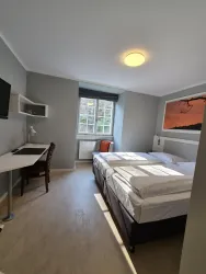 Ein modernes Hotelzimmer mit zwei Betten und einem Schreibtisch. Es gibt ein Fenster mit Tageslicht und einen Fernseher an der Wand.