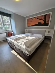 Ein helles Schlafzimmer mit zwei Einzelbetten und einem großen Fenster. An der Wand hängt ein Kunstwerk mit einem Abendhimmel.