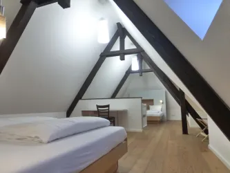 Ein modernes Schlafzimmer im Dachgeschoss mit Holzbalken und einem großen Fenster. Die Einrichtung ist einfach und gemütlich, ideal für einen entspannenden Aufenthalt.