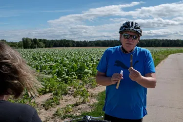 Ein Radfahrer in einem blauen T-Shirt steht neben einem Feld. Er hält einen Stift in der Hand und blickt in die Kamera.