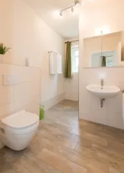 Ein modernes Badezimmer mit hellen Fliesen und minimalistischer Ausstattung. Es gibt eine Toilette, ein Waschbecken und ein Fenster mit grünen Vorhängen.