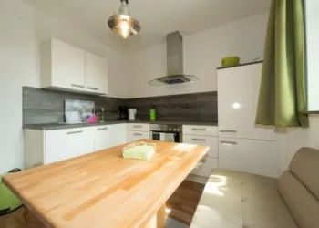 Eine moderne Küche mit weißen Möbeln und Holzdetails. Der Tisch ist in der Mitte, und es gibt eine gemütliche Sitzgelegenheit.