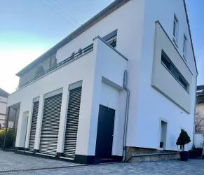 Ein modernes, weißes Haus mit klaren Linien und großen Fenstern. Der Eingangsbereich ist einfach gestaltet, mit einem schwarzen Eingangstor und stilvoller Fassadengestaltung.