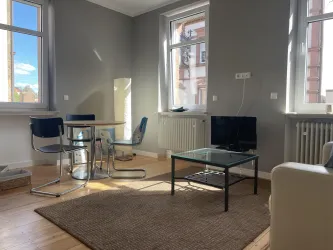 Ein helles Wohnzimmer mit einem Esstisch, drei Stühlen und einem Fernseher. Große Fenster lassen viel Licht herein, und der Holzfußboden sorgt für eine warme Atmosphäre.