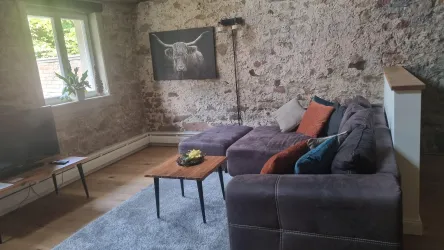 Ein gemütliches Wohnzimmer mit einer Steinfassade und Holzdeckenbalken. An der Wand hängt ein Bild von einem Rind, und es gibt eine bequeme Couch mit Kissen.