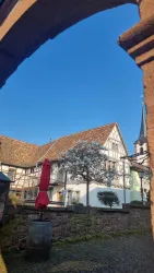 Ein malerischer Innenhof mit traditionellen Fachwerkhäusern im Hintergrund. Ein roter Sonnenschirm steht in der Mitte, unter einem blauen Himmel.