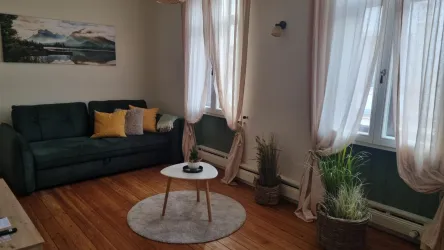 Ein gemütliches Wohnzimmer mit einem grünen Sofa und bunten Kissen. Der Raum hat große Fenster mit Vorhängen und einen runden Teppich auf dem Holzboden.