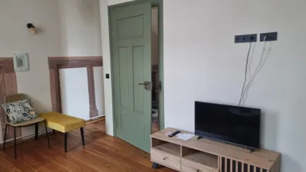 Ein modern eingerichteter Raum mit einem Fernseher auf einem Holzschrank. An der Wand befindet sich eine grüne Tür und ein Stuhl mit einem kuscheligen Kissen.