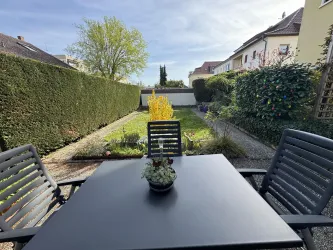 Ein gepflegter Garten mit grünen Hecken und bunten Pflanzen. Ein schwarzer Tisch mit Stühlen lädt zum Verweilen ein.